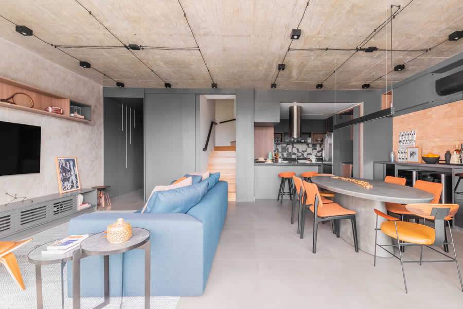 A primazia da est&eacute;tica industrial desse projeto assinado pelo arquiteto Bruno Moraes, assinala a contemporaneidade das cores pontuais por meio do sof&aacute; azul e as cadeiras laranja da sala de jantar | Foto: Guilherme Pucci