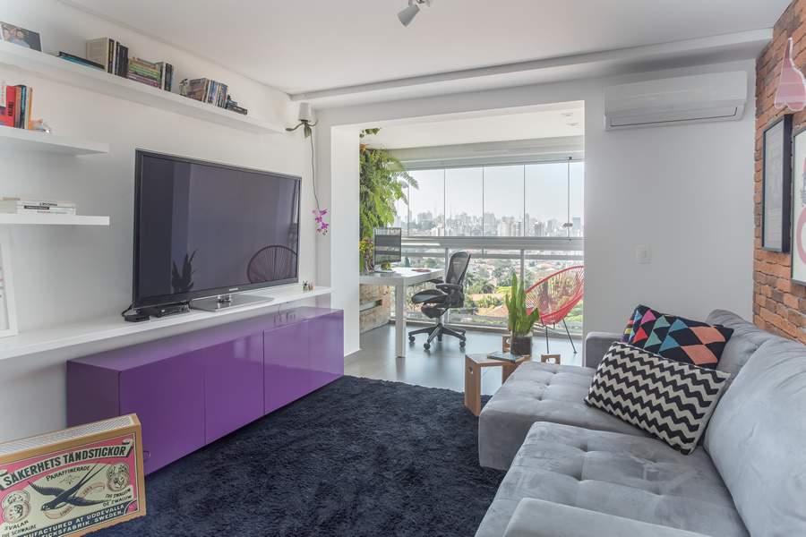  Na sala de estar integrada com a varanda, as arquitetas Ieda e Carina Korman, do Korman Arquitetos, investiram em um rack no tom Very Peri, que trouxe a contemporaneidade buscada para a morada do jovem casal. A TV foi apoiada em uma prateleira, instalada de ponta a ponta na parede | Foto: JP Image